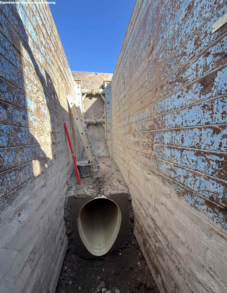 Narrow construction trench between weathered concrete walls with a large cylindrical pipe resting in muddy ground; a ladder and tools are nearby, blue sky above.
