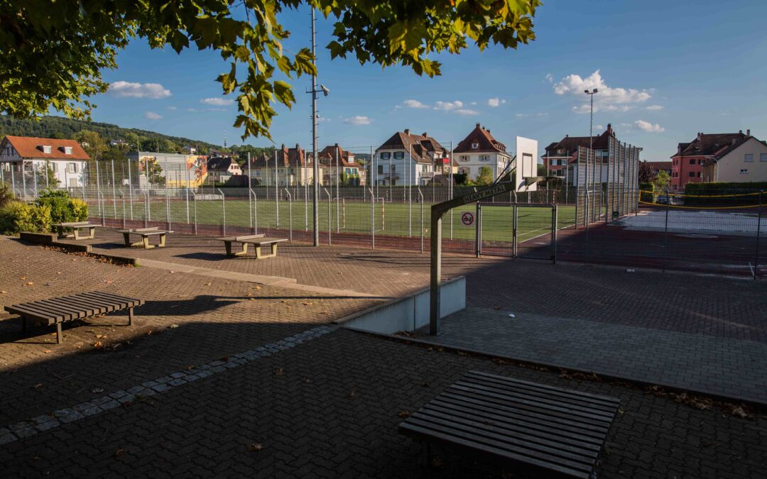 Basketball court behind a tall metal fence with benches in a sunny urban park and residential buildings in the background under a blue sky with trees overhead