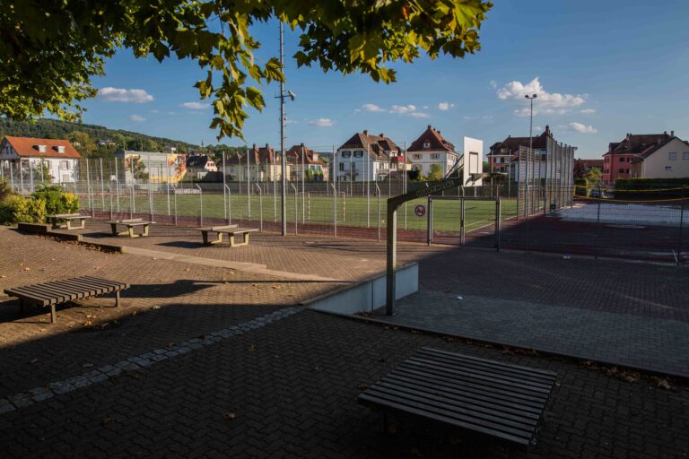 Basketball court behind a tall metal fence with benches in a sunny urban park and residential buildings in the background under a blue sky with trees overhead