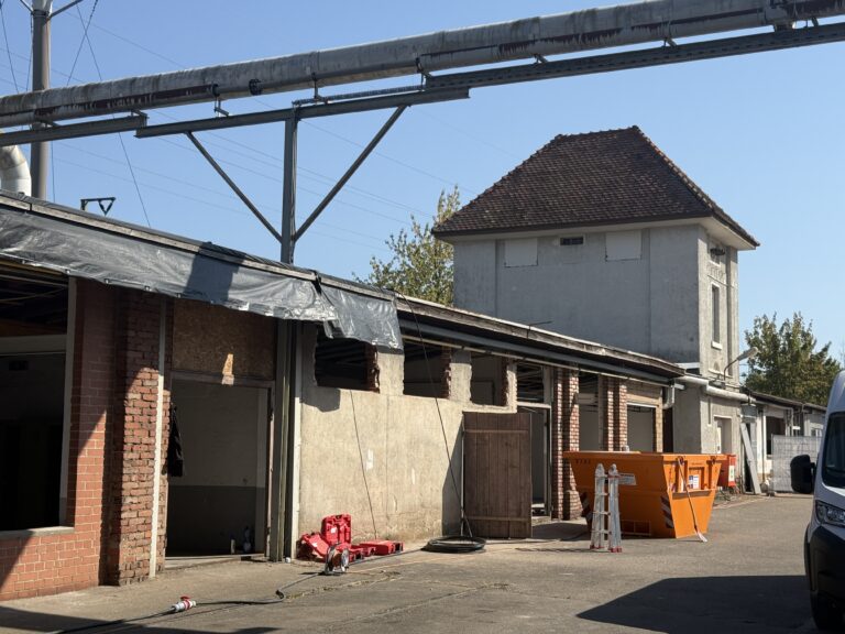 Industrial building with overhead metal pipes and scaffolding, construction barriers, and hoses on a sunny day