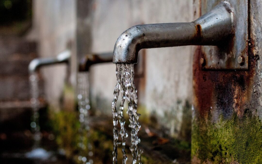 Rusty outdoor faucet with water streaming from the spout onto a mossy wall.