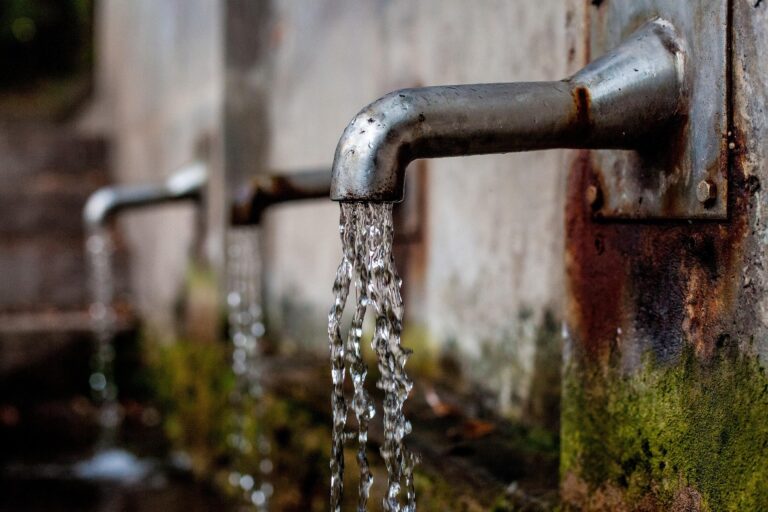 Rusty outdoor faucet with water streaming from the spout onto a mossy wall.
