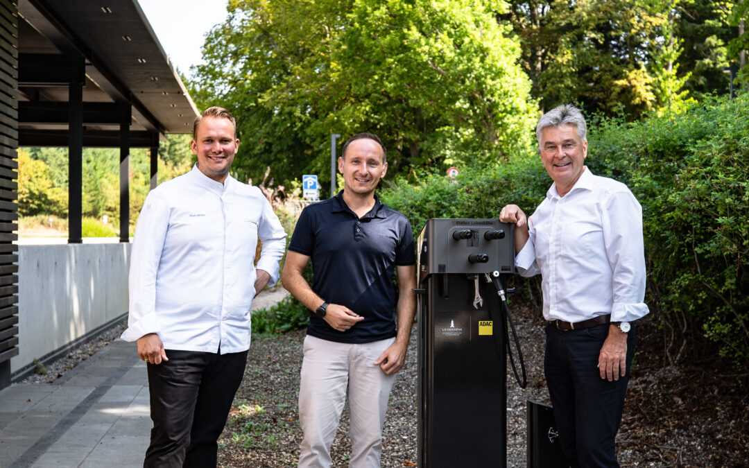 Three men stand outdoors beside a tall black charging station, smiling at the camera.