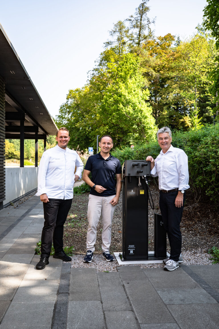 Three men stand outdoors beside a tall black charging station, smiling at the camera.