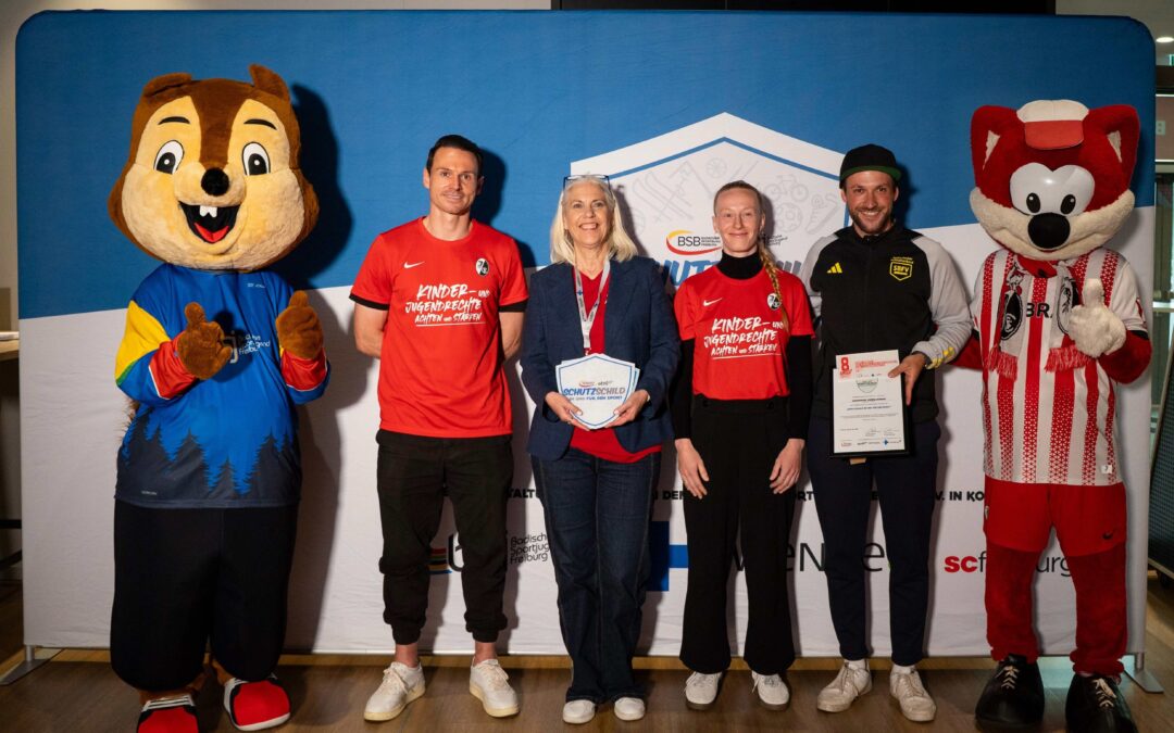 Group of five people, plus two costumed mascots, posing with certificates at an indoor event backdrop.