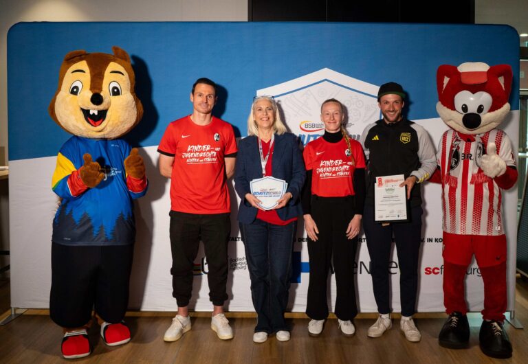 Group of five people, plus two costumed mascots, posing with certificates at an indoor event backdrop.