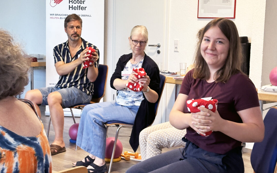 People seated in a room, a smiling young woman in a maroon shirt holds a red polka-dotted cloth; a banner with 'Deutscher Roter Helfer' is in the background.