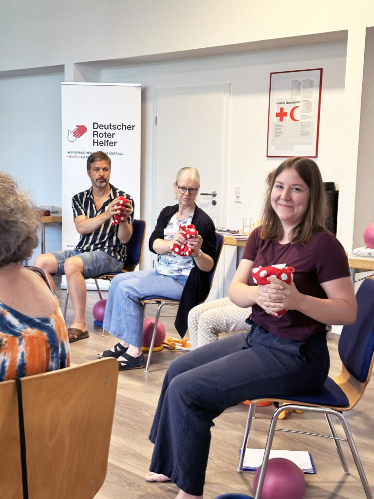 People seated in a room, a smiling young woman in a maroon shirt holds a red polka-dotted cloth; a banner with 'Deutscher Roter Helfer' is in the background.
