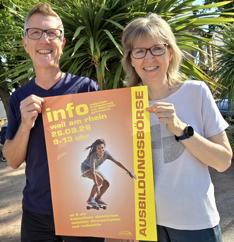 Two smiling adults wearing glasses hold an orange poster outdoors, with a large yellow stripe and palm plants in the background.