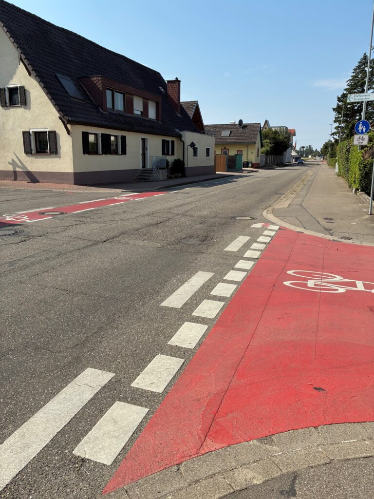 Residential street scene with a red bike lane on the right and white crosswalk markings; beige houses with shutters line the left sidewalk under a blue sky.
