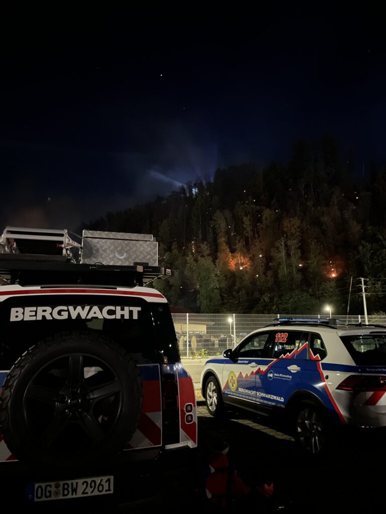 Nighttime view of emergency vehicles in a parking area, with a forest fire burning on the hillside in the background and blue lights nearby.