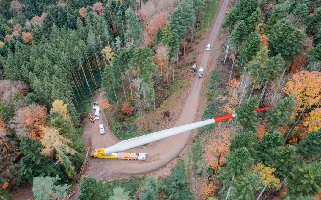 Aerial view of a forested area with a winding dirt road; a large white wind turbine blade sits on a yellow trailer as several vehicles pass by among autumn trees.