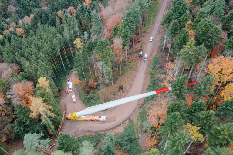 Aerial view of a forested area with a winding dirt road; a large white wind turbine blade sits on a yellow trailer as several vehicles pass by among autumn trees.