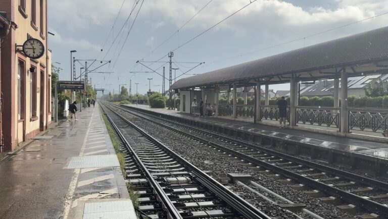 Train station platform with tracks between two platforms, a pink building with a round clock on the left, and people waiting under a long curved shelter on the right. Wet ground from recent rain.