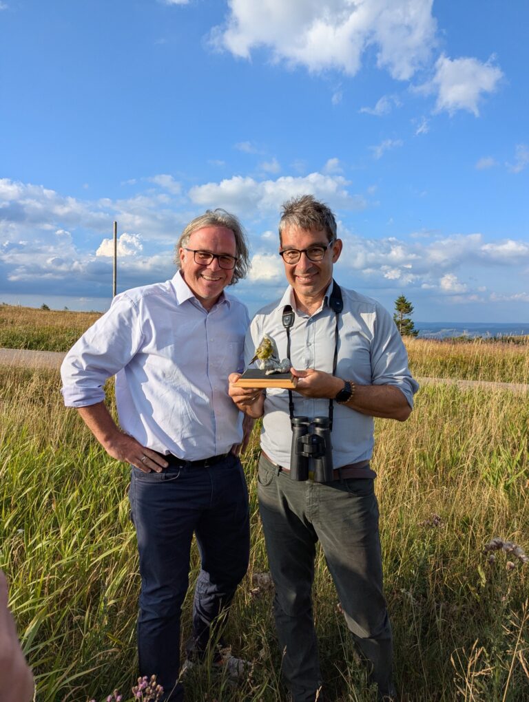 Two men standing in a grassy field smiling and holding a small bird trophy together against a blue sky.