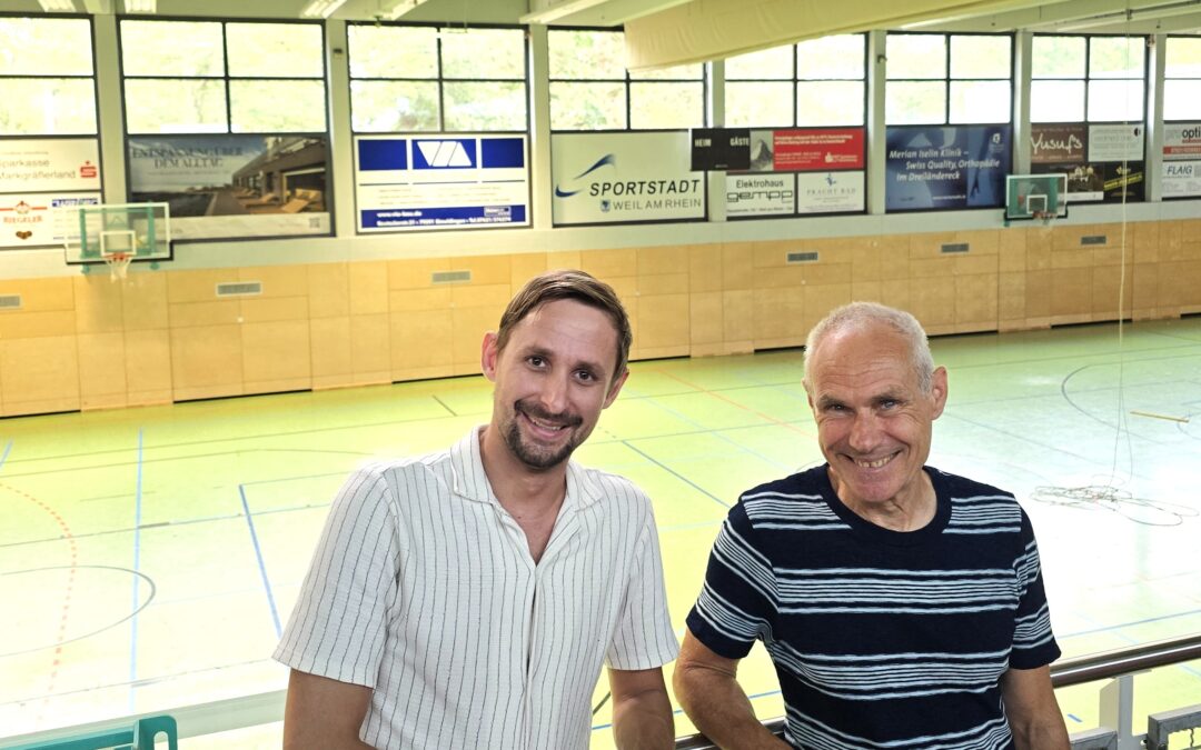 Two men pose at a railing inside a basketball gym, smiling with a court and boards behind them.
