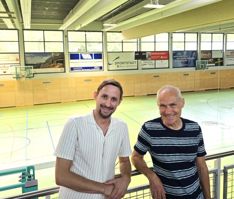 Two men pose at a railing inside a basketball gym, smiling with a court and boards behind them.