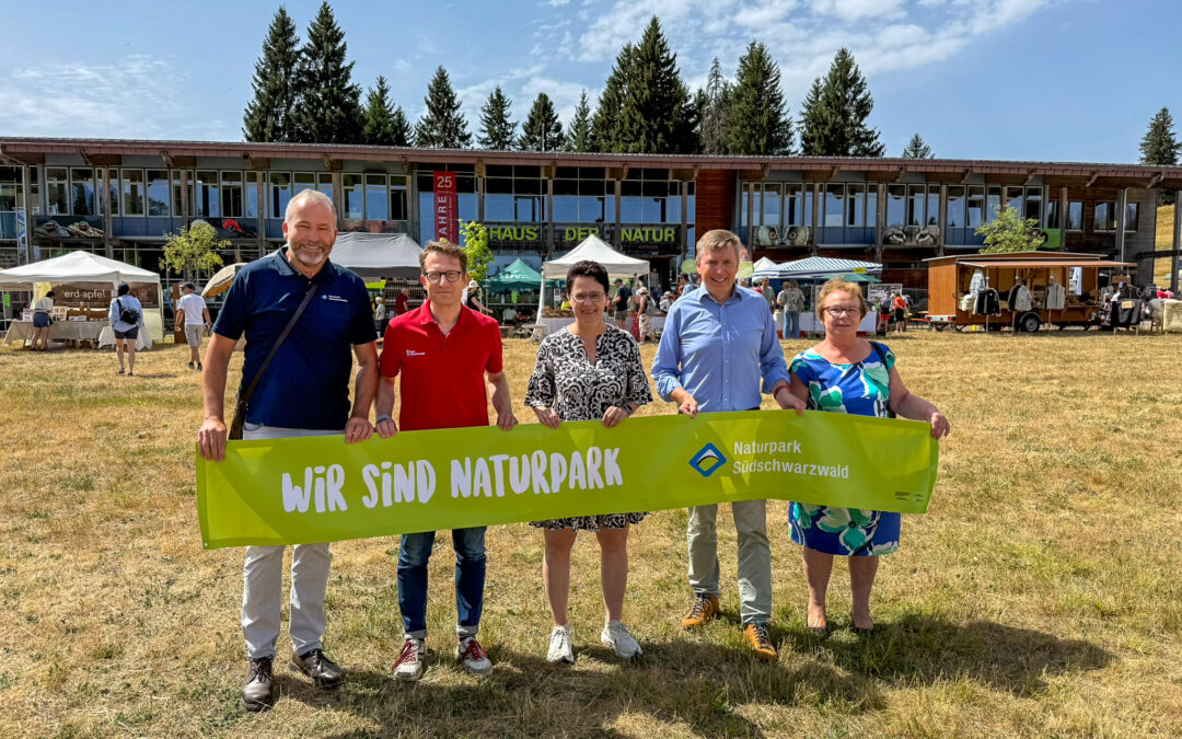 Five adults posing with a bright green banner that reads 'Wir sind Naturpark' at an outdoor nature park event, with market stalls behind them.