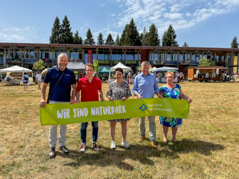 Five adults posing with a bright green banner that reads 'Wir sind Naturpark' at an outdoor nature park event, with market stalls behind them.