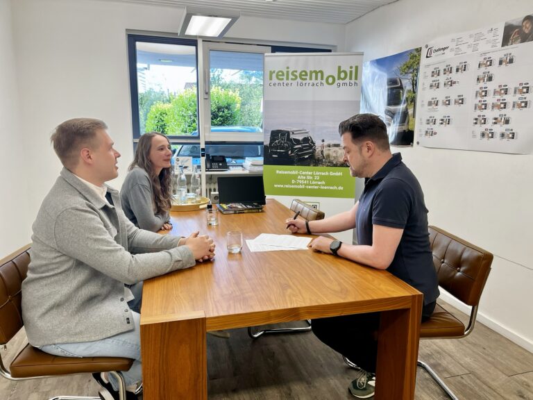 Three people sit around a wooden table in a bright office, with a man on the right signing papers as two colleagues look on.