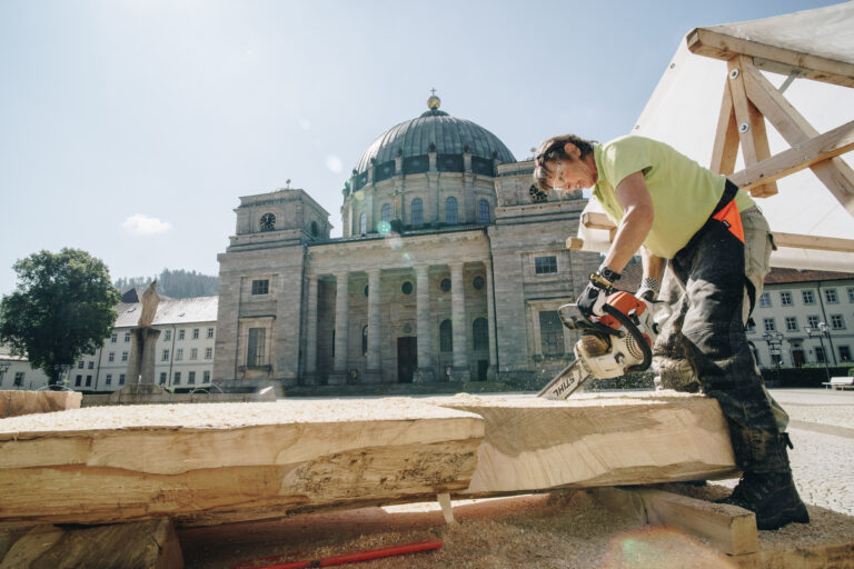 Construction worker cutting a wooden beam with a power saw in front of a historic cathedral.
