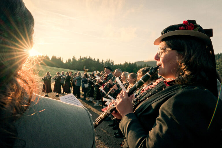 Outdoor brass band performance at sunset, viewed from the audience side with players in uniform and sun flare in the foreground.