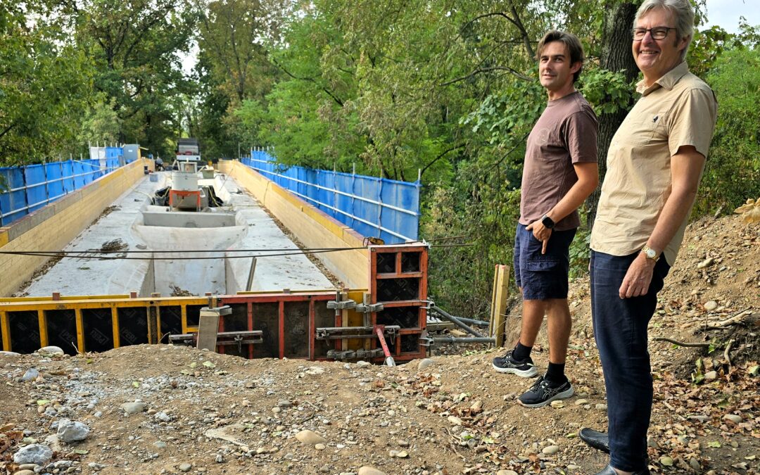 Two men stand at the edge of a construction site beside a deep, partially built concrete trench, with blue safety barriers and trees in the background.