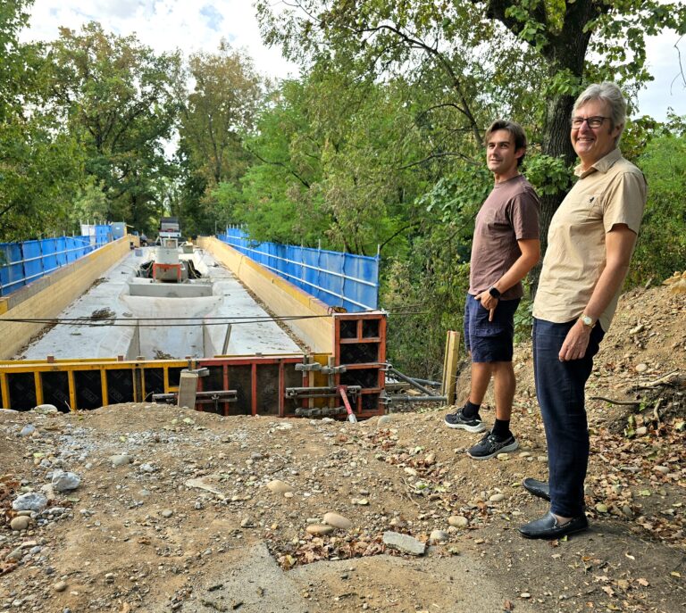 Two men stand at the edge of a construction site beside a deep, partially built concrete trench, with blue safety barriers and trees in the background.