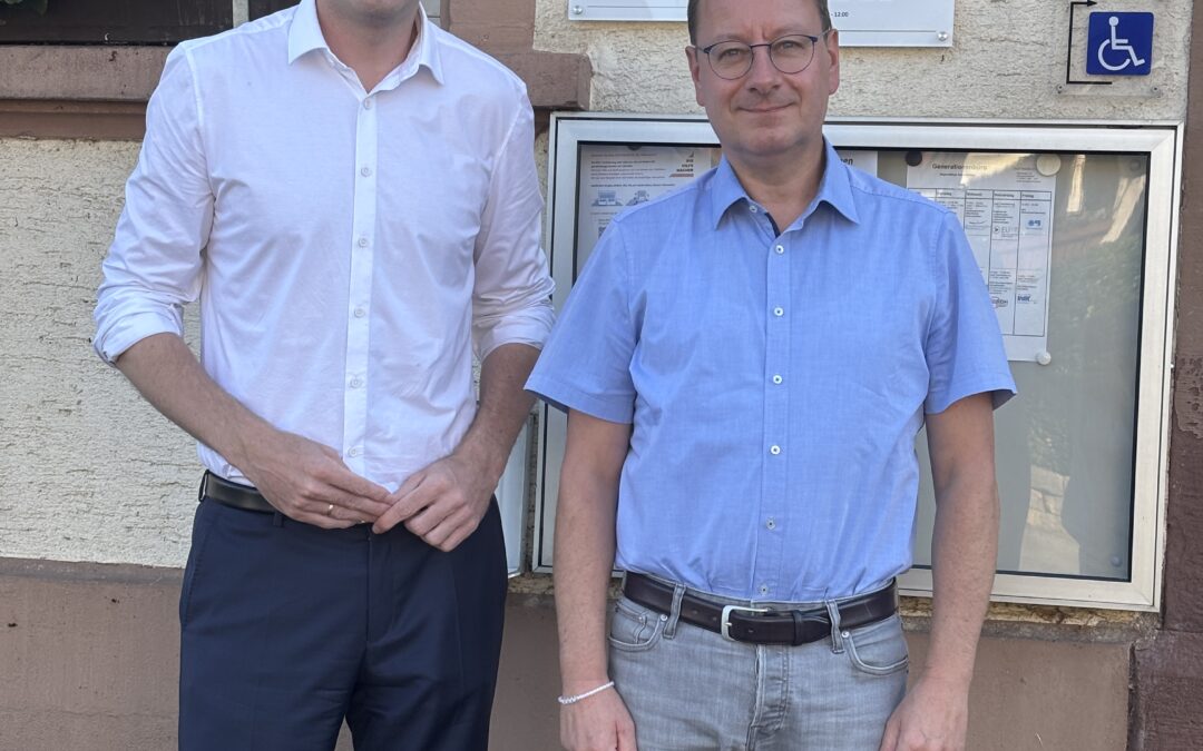 Two men in light shirts pose outside a municipal building with a sign that reads Bürgermeisteramt Gutach i.Br. Landkreis Emmendingen, smiling at the camera.