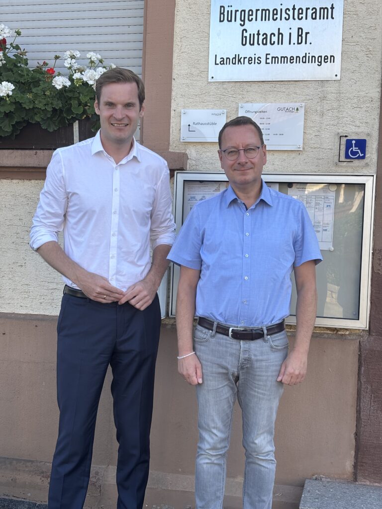 Two men in light shirts pose outside a municipal building with a sign that reads Bürgermeisteramt Gutach i.Br. Landkreis Emmendingen, smiling at the camera.
