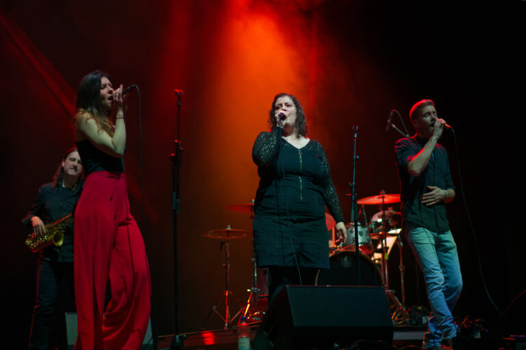 Band on stage performing live under red lights; center female singer with others around—saxophonist on left and male vocalist on right.
