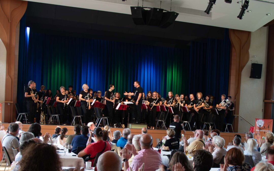 A community band on stage, conductor directing, as audience sits at tables and applauds in the foreground.