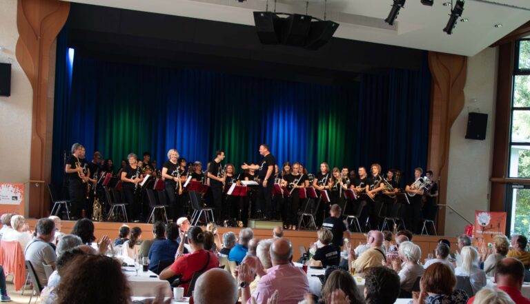 A community band on stage, conductor directing, as audience sits at tables and applauds in the foreground.