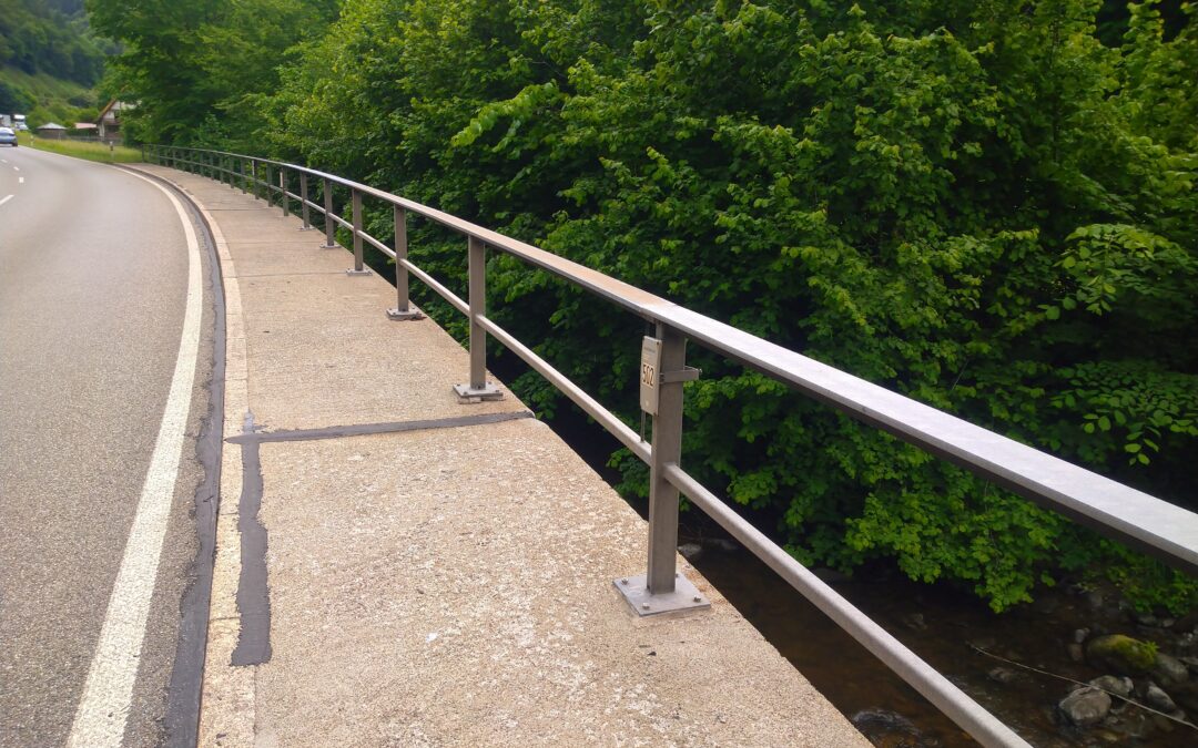 Curved road and sidewalk on a bridge with metal railing, dense green trees on the right and a river below the gap between rails.