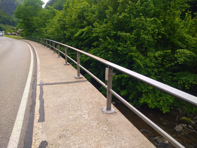 Curved road and sidewalk on a bridge with metal railing, dense green trees on the right and a river below the gap between rails.