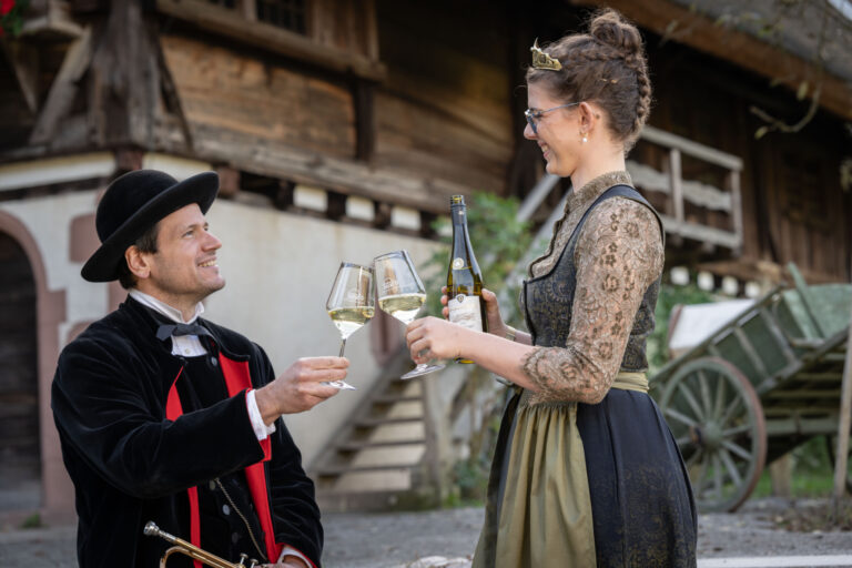 Two costumed adults toast with wine outdoors: a man in a velvet black outfit and hat and a woman in a lace dress holding a wine bottle and glasses.