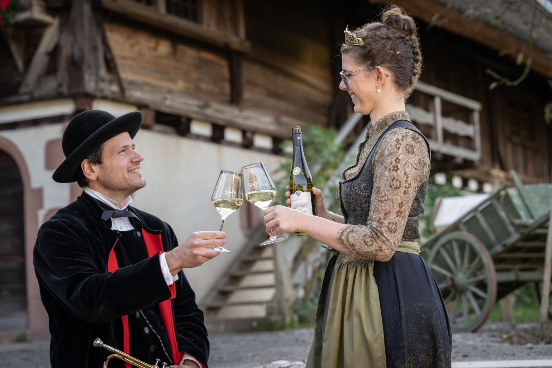 Two costumed adults toast with wine outdoors: a man in a velvet black outfit and hat and a woman in a lace dress holding a wine bottle and glasses.
