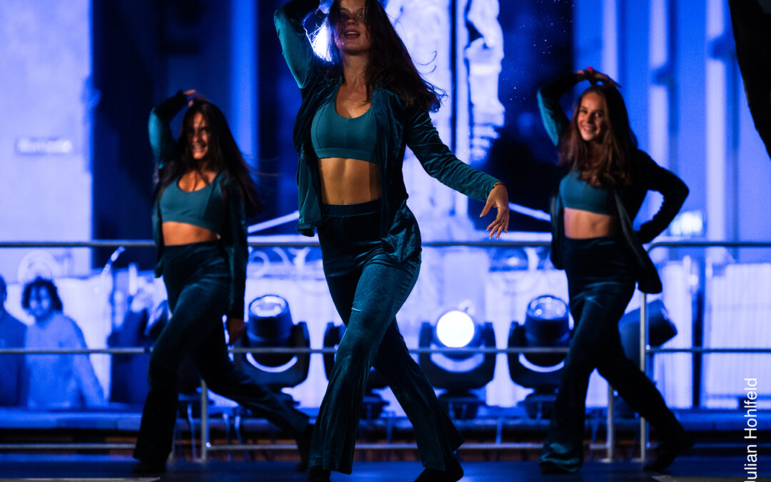 Three women in teal outfits dance on a blue-lit stage, mid-performance with energetic motions.