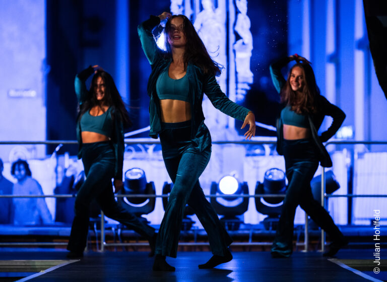 Three women in teal outfits dance on a blue-lit stage, mid-performance with energetic motions.