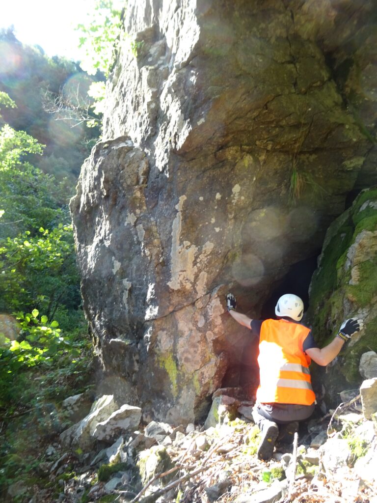 Climber wearing a white helmet and bright orange safety vest grips a rocky cliff face outdoors.