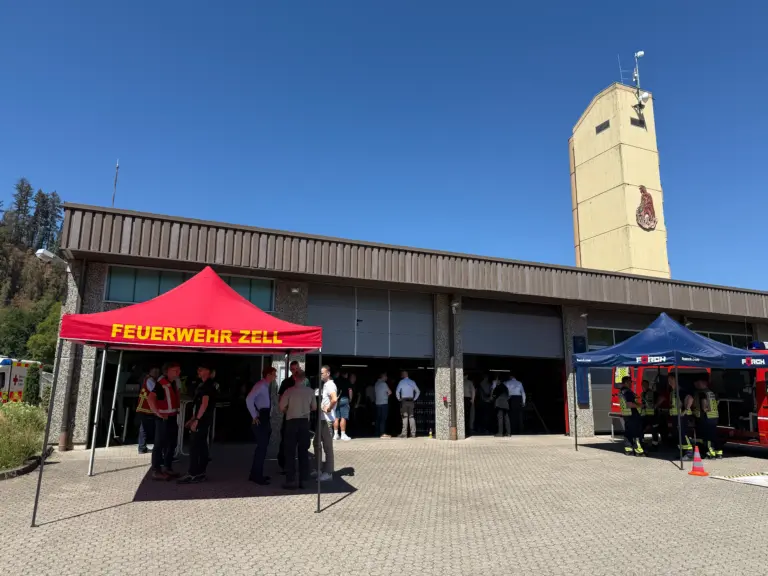 Outdoor firefighting event under red tent reading 'FEUERWEHR ZELL' with people and firefighters gathered outside a building.