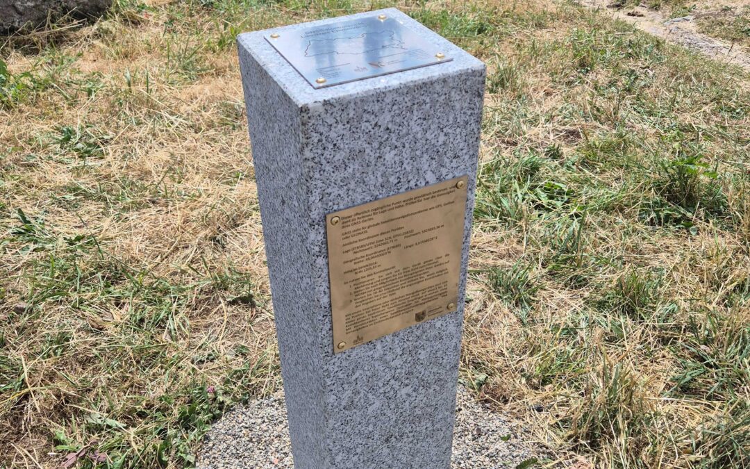 Granite pedestal with a bronze plaque in a grassy, rocky park area; wildflowers and a signpost visible in the background.