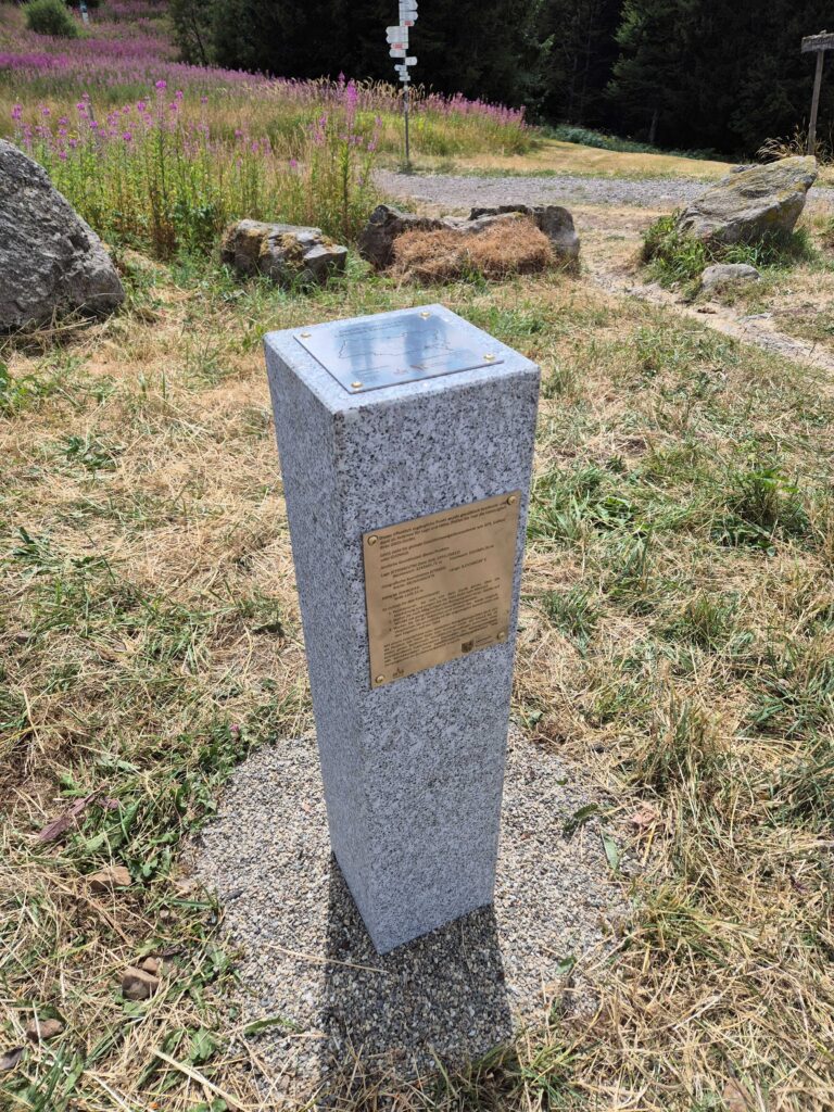 Granite pedestal with a bronze plaque in a grassy, rocky park area; wildflowers and a signpost visible in the background.
