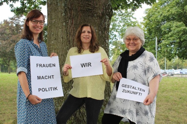 Three women stand by a tree outdoors, holding signs that read 'Frauen Macht Politik', 'Wir', and 'Gestalten Zukunft!'