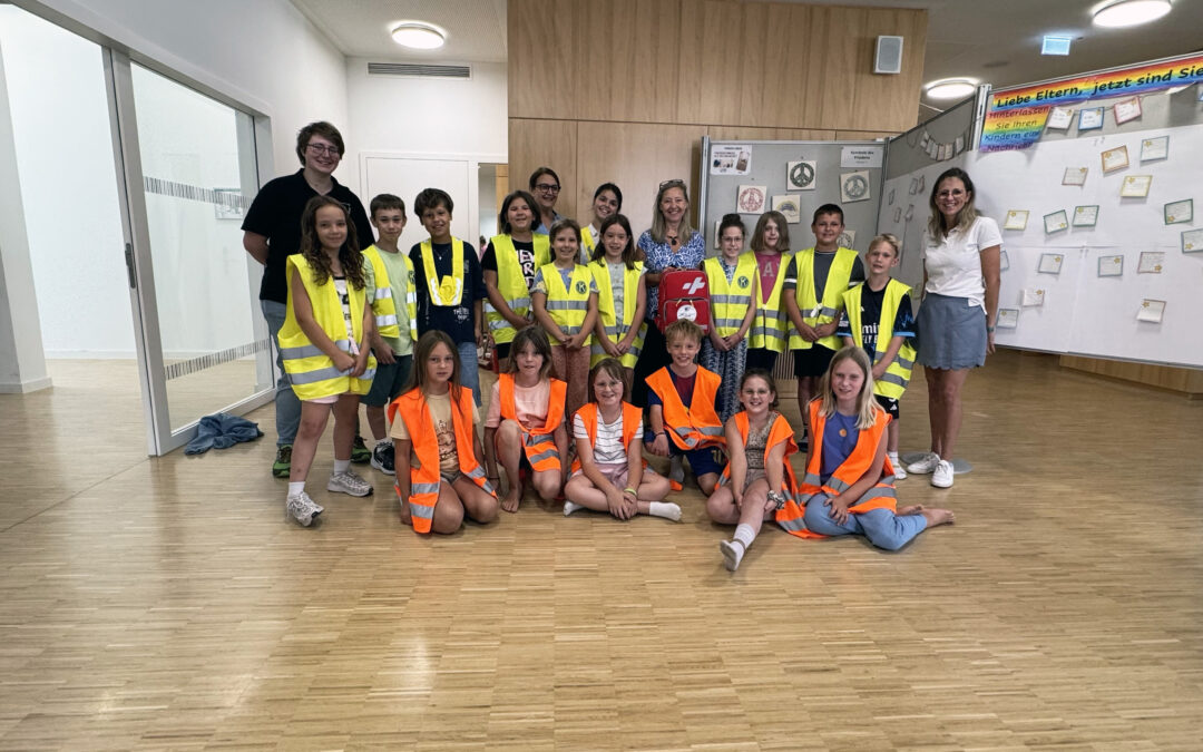 Group of children wearing yellow and orange high-visibility vests posing for a photo in a hallway.