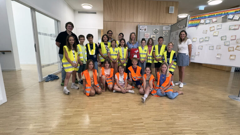 Group of children wearing yellow and orange high-visibility vests posing for a photo in a hallway.