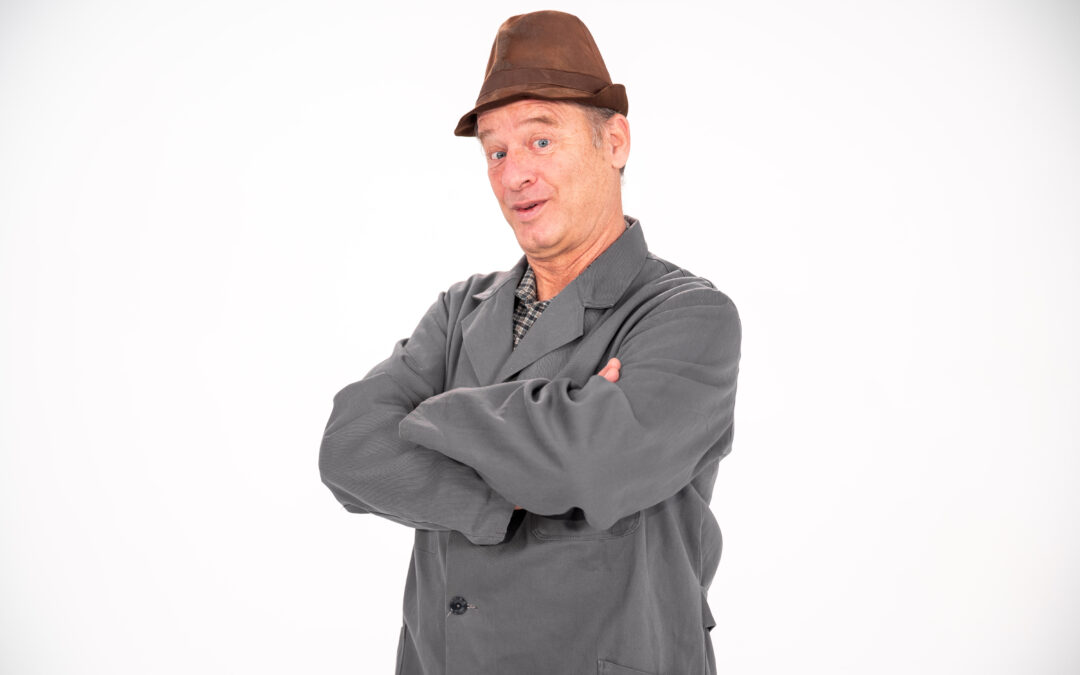 Man in gray work coat with arms crossed, wearing a brown fedora, posing against a white background; looks confident or playful.
