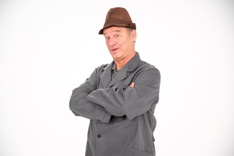 Man in gray work coat with arms crossed, wearing a brown fedora, posing against a white background; looks confident or playful.
