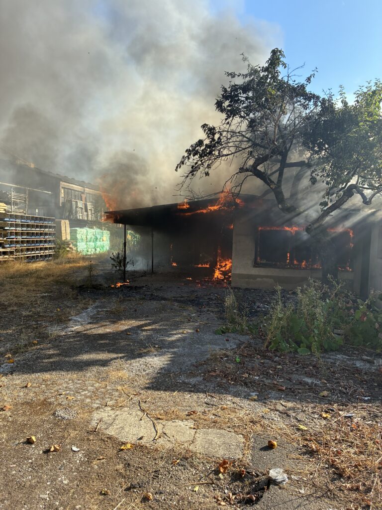 A small building is on fire with flames along the roof and a large plume of smoke rising, while a tree stands nearby and pallets are stacked to the left in an outdoor yard.