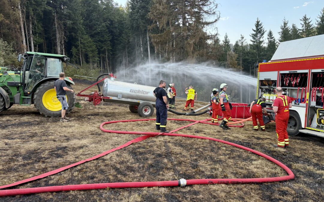 Firefighters connect hoses to a water tanker in a forest clearing; a red fire truck is visible on the right side.
